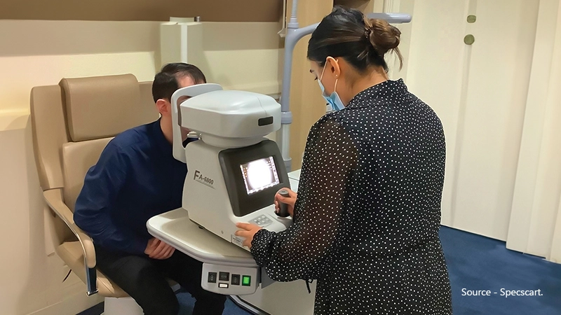 An eye doctor operates an auto-refractor machine (FA-6000) on a male patient to objectively measure his refractive error, including sphere, cylinder, and axis.