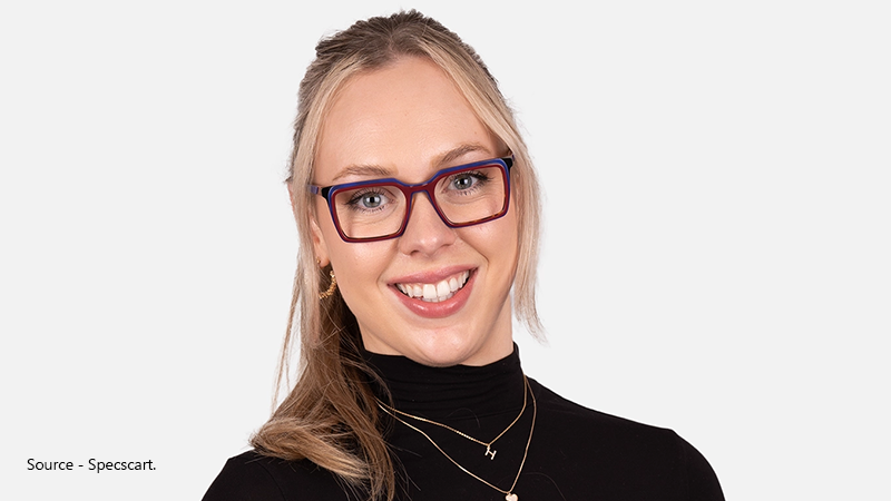 A smiling woman with blonde hair wears bold, rectangular eyeglasses with burgundy and blue frames, representing the stylish correction of astigmatism.