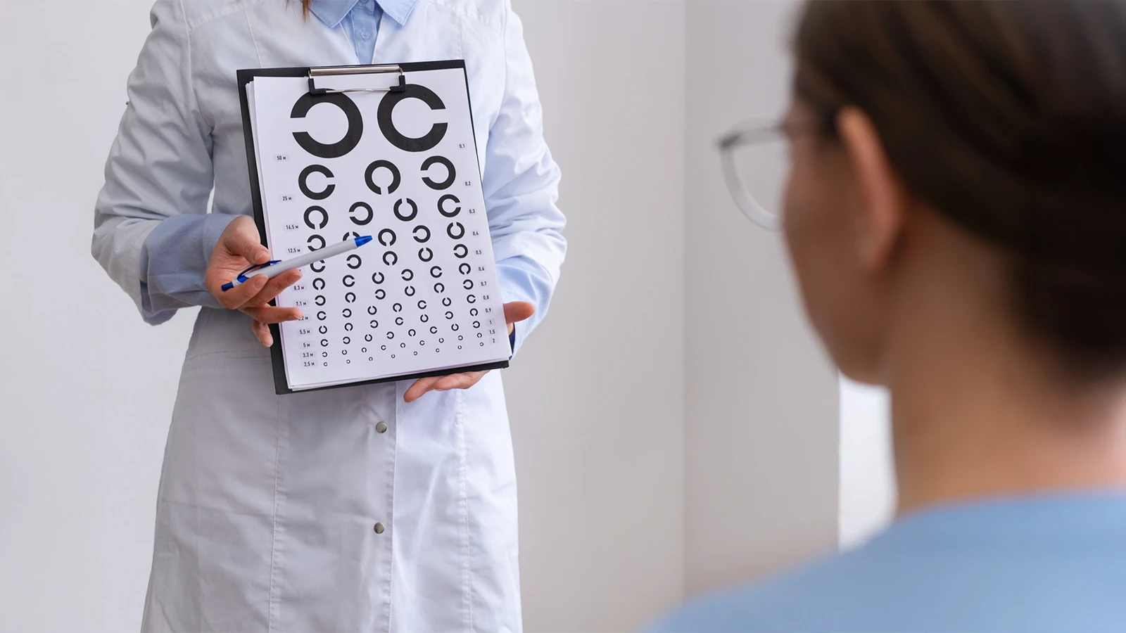 An optometrist in a white coat holding a Landolt C eye chart, pointing to the rings while performing a visual acuity test for a patient.