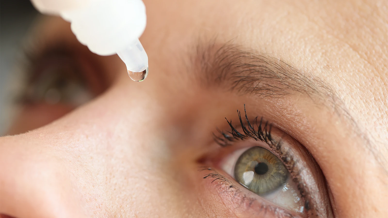 Close-up of a light green eye with an eye drop falling from a white dropper bottle tip.