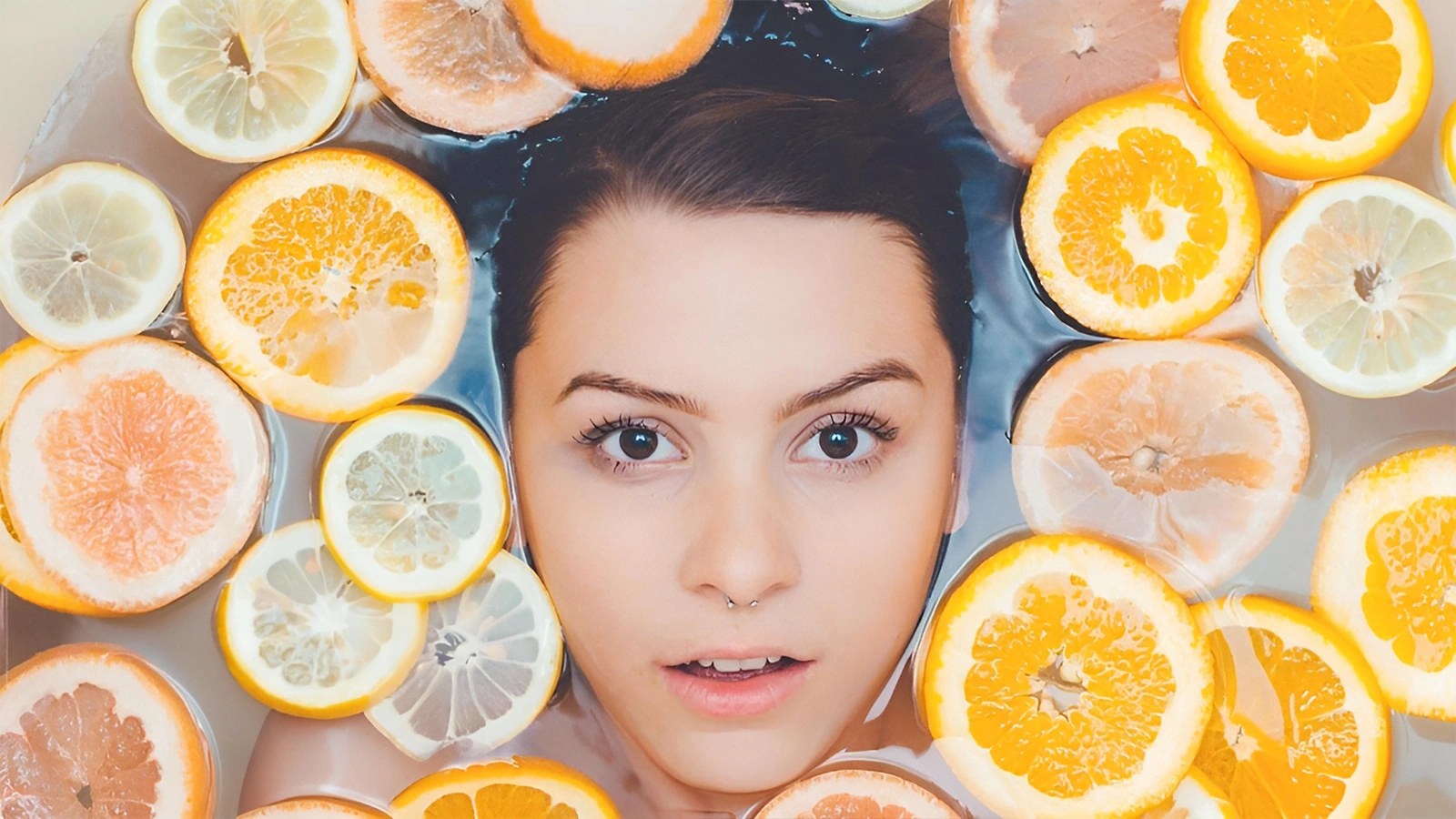 A woman's face submerged in water, surrounded by floating slices of oranges, lemons, and grapefruits, looking directly at the camera.