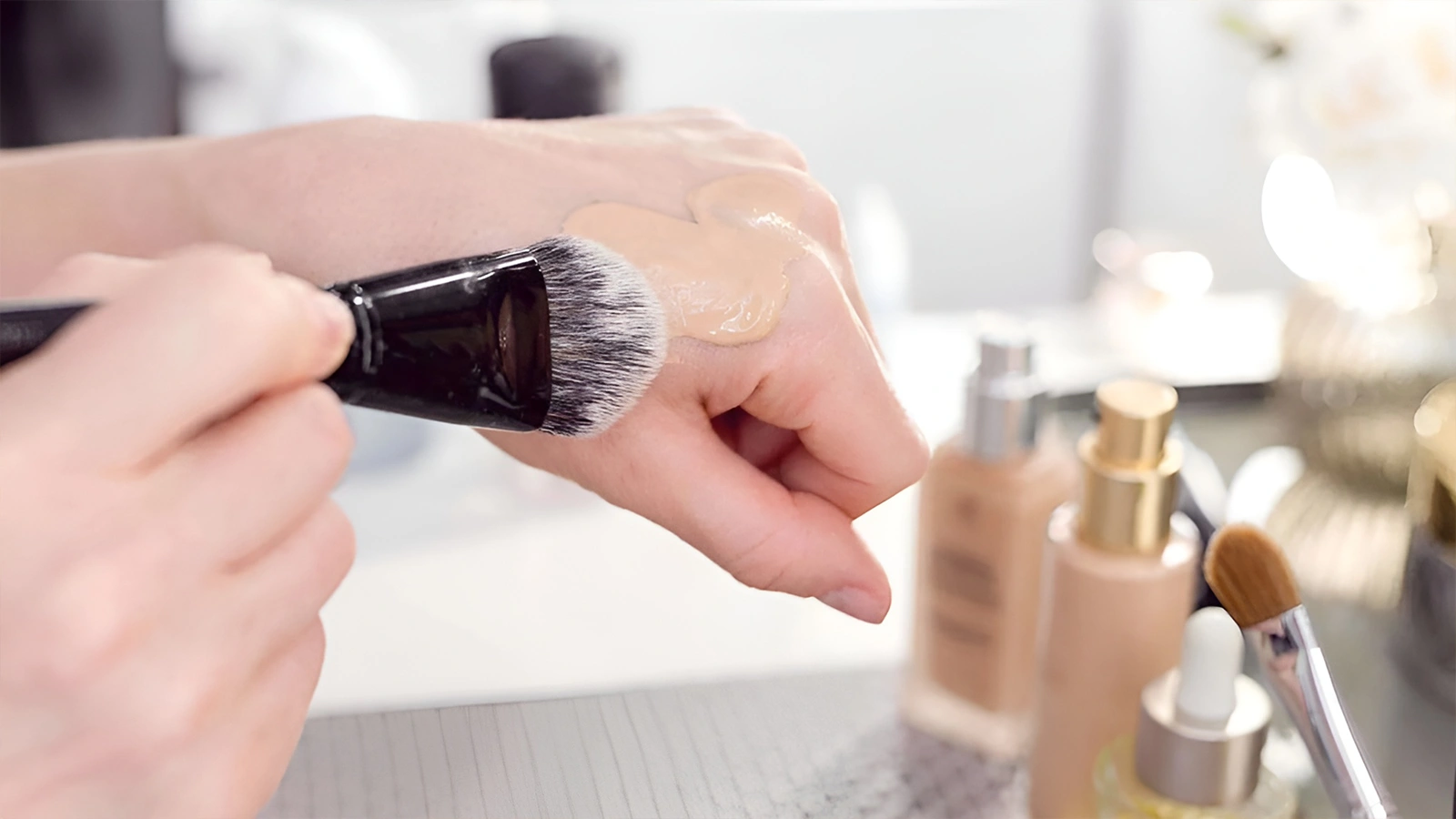 Close-up of a person using a makeup brush to apply liquid foundation to their hand, demonstrating how to use concealer for under-eye discoloration.
